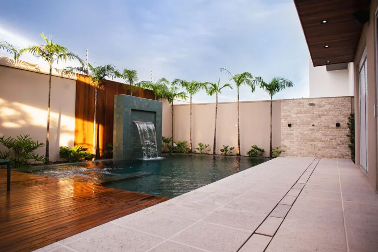 Área de piscina com cascata, deck de madeira e piso claro no entorno